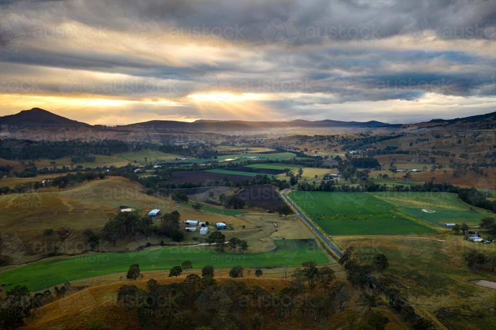 Sun rays piercing through the cloudy sky over farmland rimmed in hills - Australian Stock Image