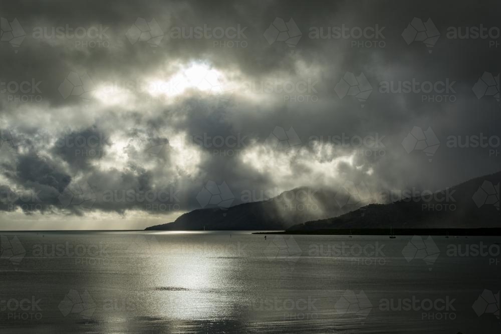 Sun shining through the clouds onto ocean and mountain - Australian Stock Image