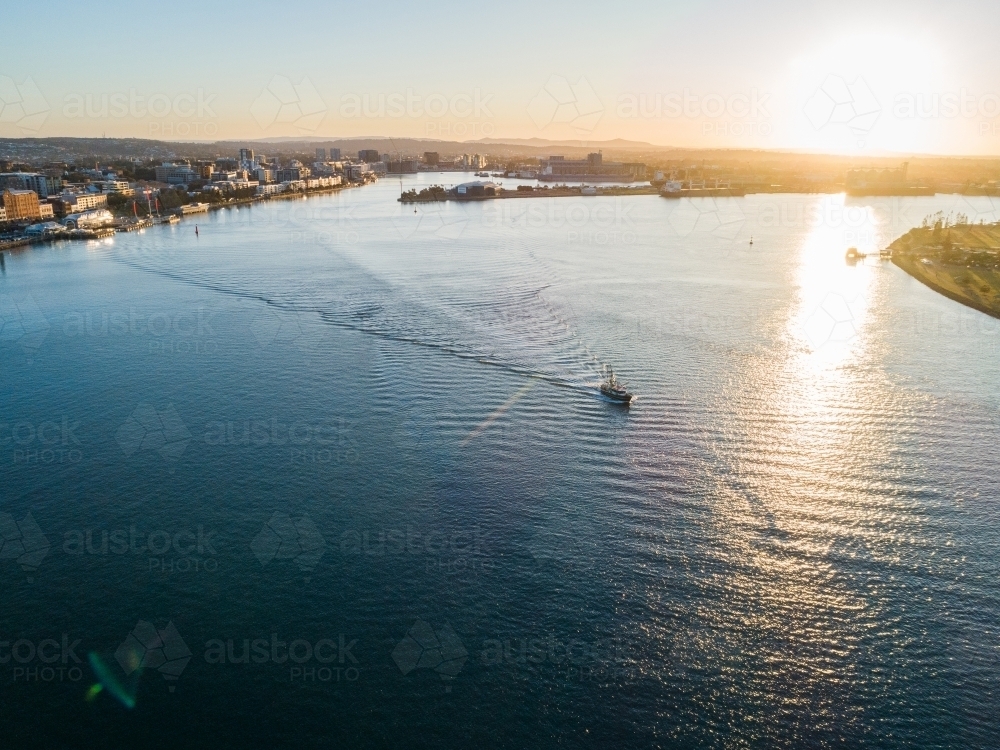 Sunset light over Newcastle Harbour with small boat leaving a wake in sparkling blue water - Australian Stock Image