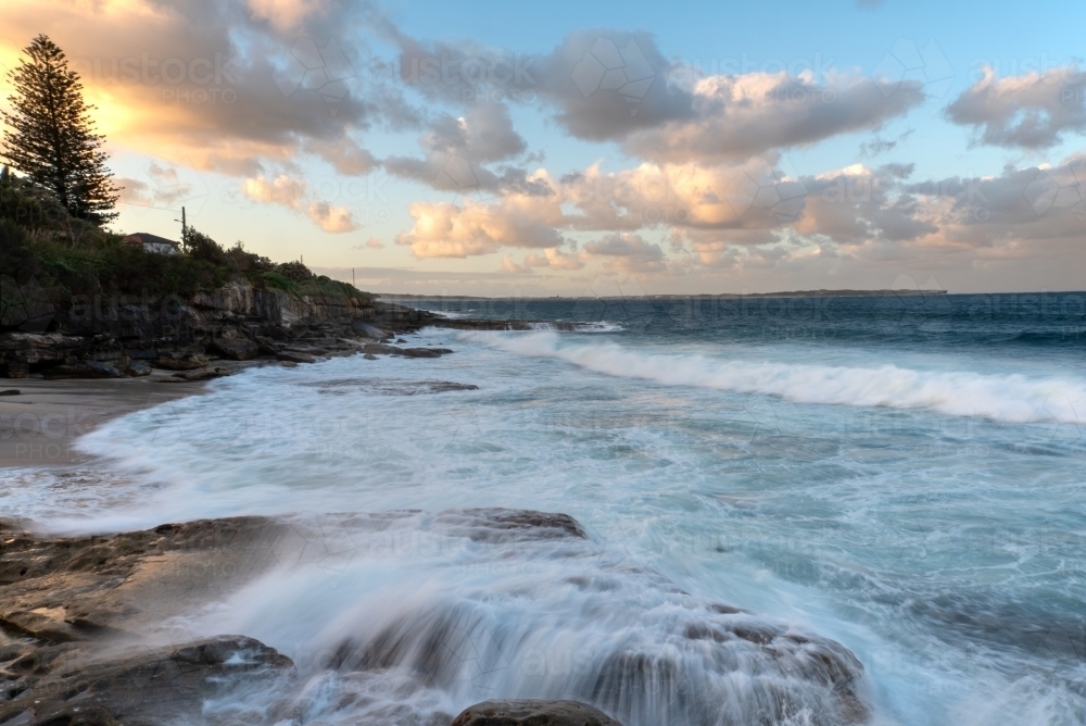 Sunset with waves crashing over rocks in a long exposure - Australian Stock Image