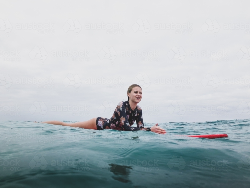 Surfing Teenage girl lying on red surfboard in ocean smiling wearing floral swimsuit on overcast day - Australian Stock Image