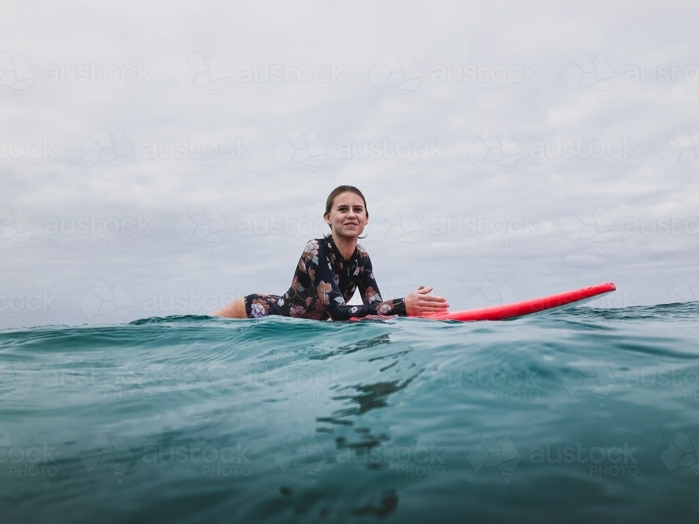 Surfing Teenage girl lying on red surfboard in ocean wearing floral swimsuit on overcast day - Australian Stock Image