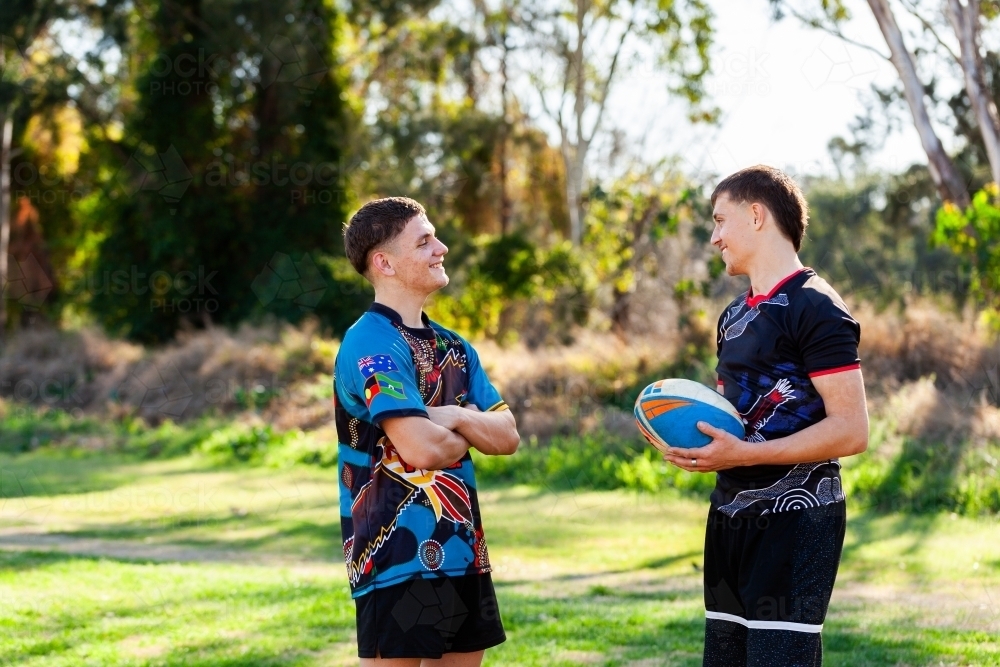 Teammates and cousins talking together on edge of sports ground holding footy - Australian Stock Image