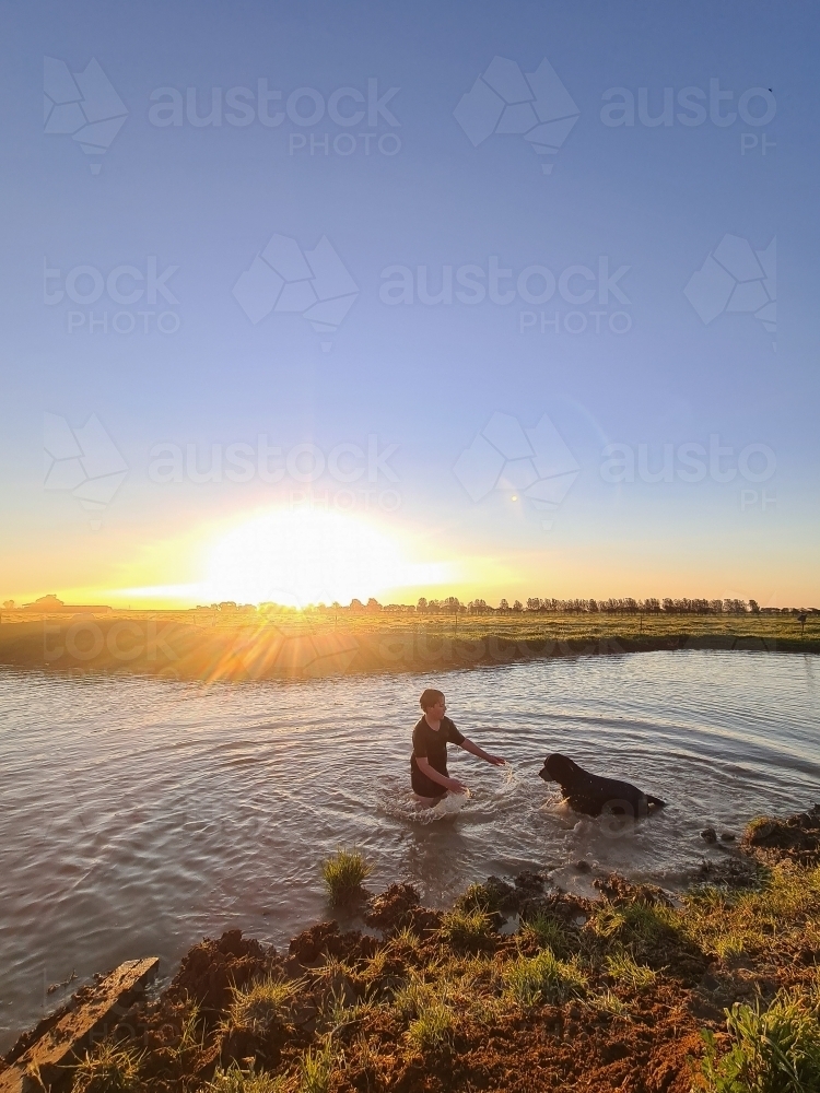 Teen boy and his dog swimming in dam at sunset. Summer life on the farm. - Australian Stock Image