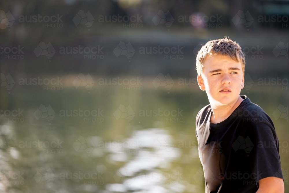 Teen boy in afternoon sunshine, looking away with water background - Australian Stock Image