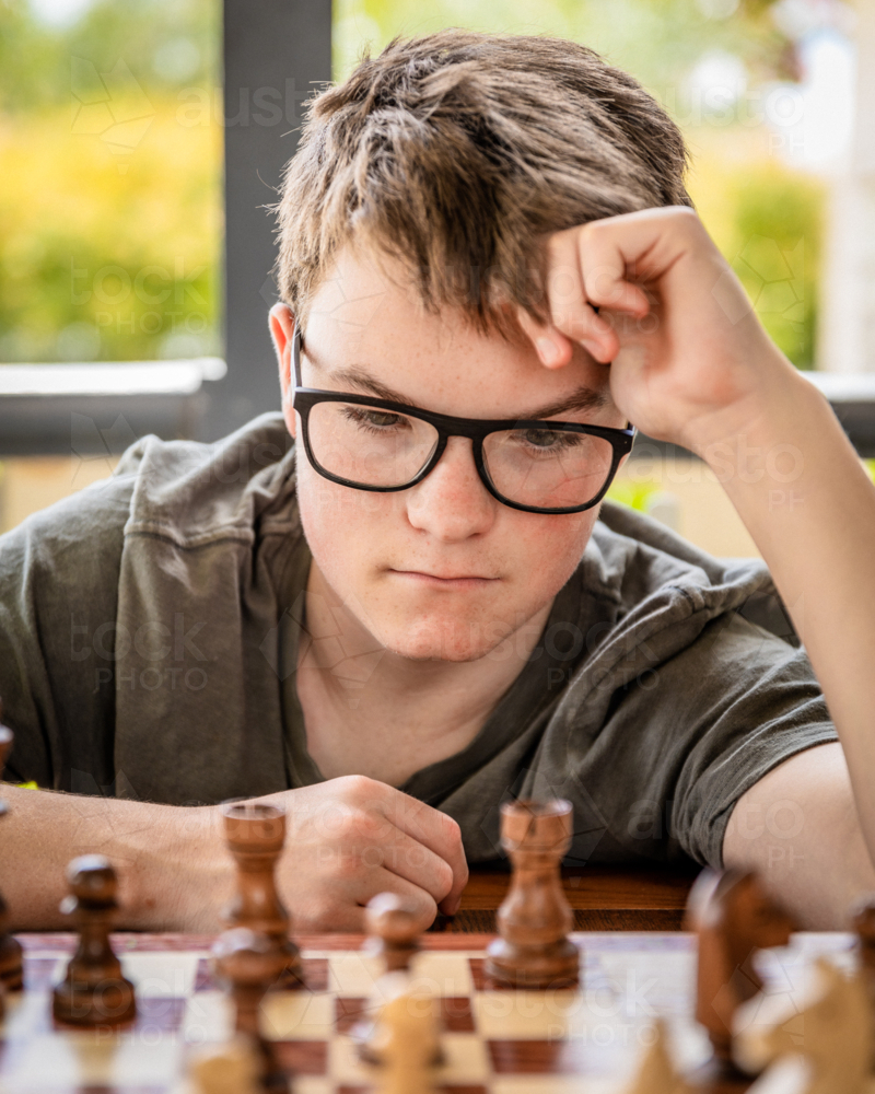 Teen boy with glasses contemplating a chess board - Australian Stock Image