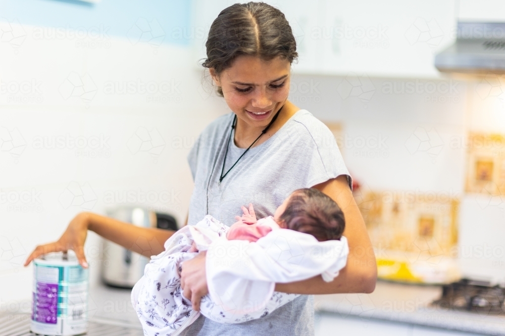 teen girl holding baby with tin of infant formula on kitchen bench - Australian Stock Image