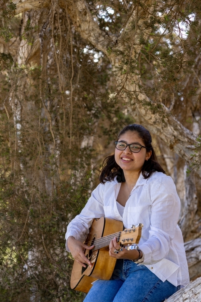 teen girl in white shirt wearing glasses strumming guitar sitting on a tree - Australian Stock Image