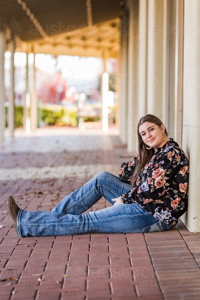 Teen girl sitting in main street smiling leaning against building - Australian Stock Image