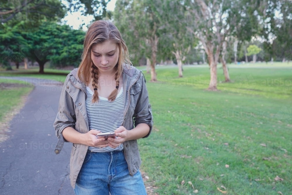 Teen girl using mobile phone while walking in the park - Australian Stock Image