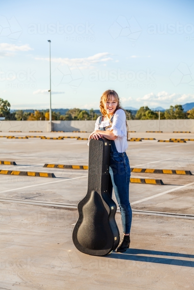 Teen musician leaning on guitar case in empty car park - Australian Stock Image