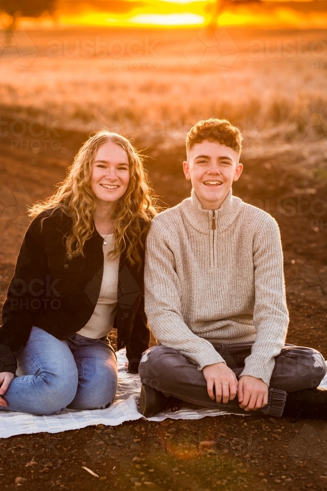 Teenage boy and girl sitting on blanket on rural dirt road at sunset - Australian Stock Image