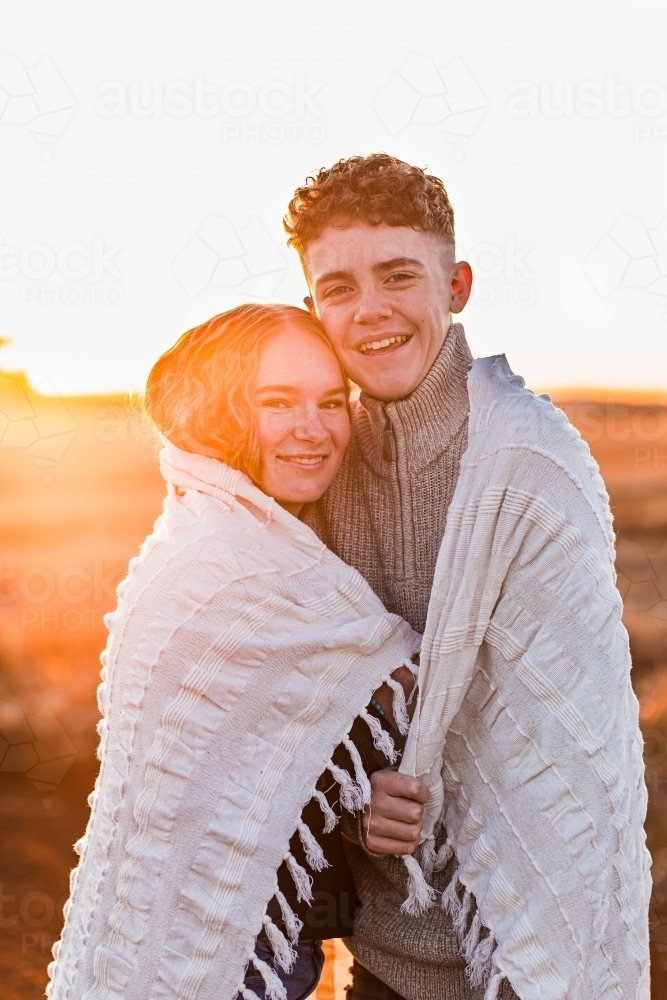 Teenage boy and girl together draped with white blanket at sunset - Australian Stock Image