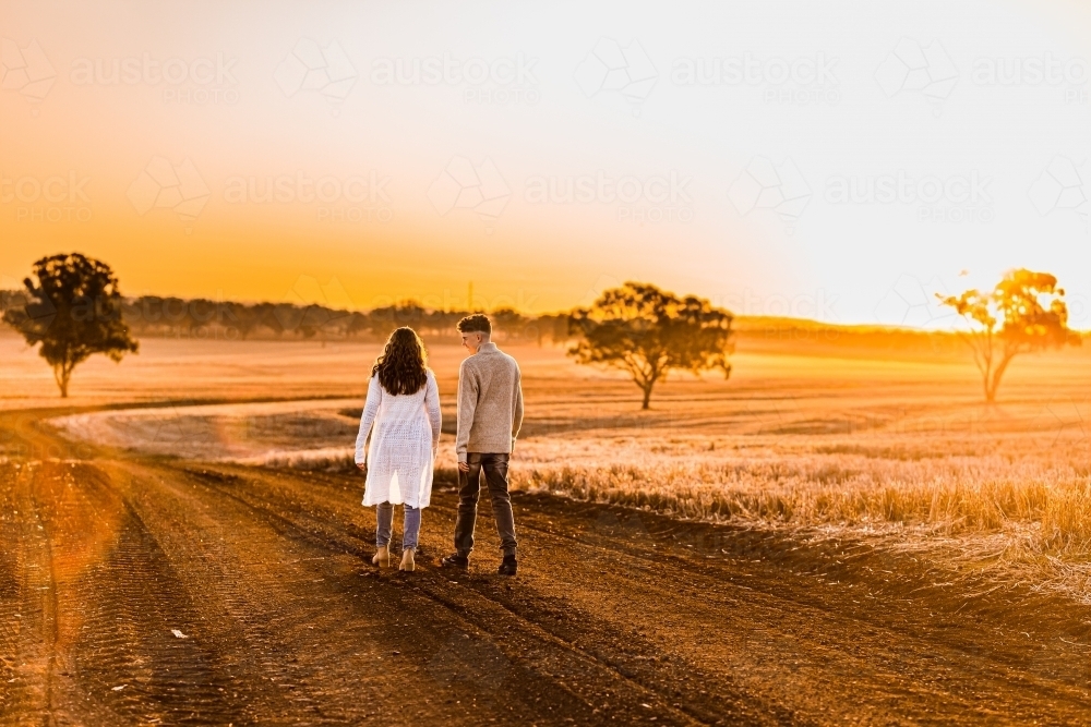 Teenage boy and girl walking together on rural dirt road at sunset - Australian Stock Image