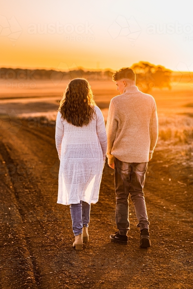 Teenage boy and girl walking together on rural dirt road at sunset - Australian Stock Image