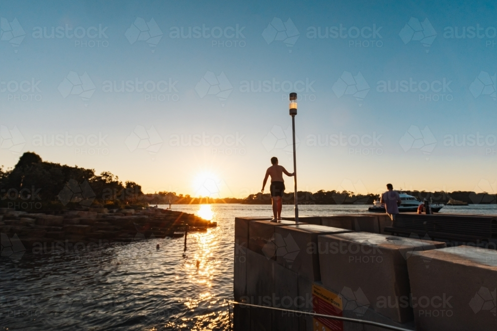 Teenage boy standing on ledge by water in bright setting sun - Australian Stock Image