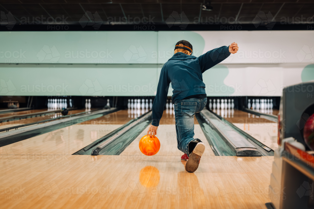 Teenage boy ten pin bowling with orange ball in brightly lit bowling alley - Australian Stock Image