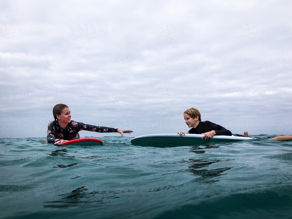 Teenage girl assisting teenage boy in the surf on longboard surfboards on overcast day - Australian Stock Image