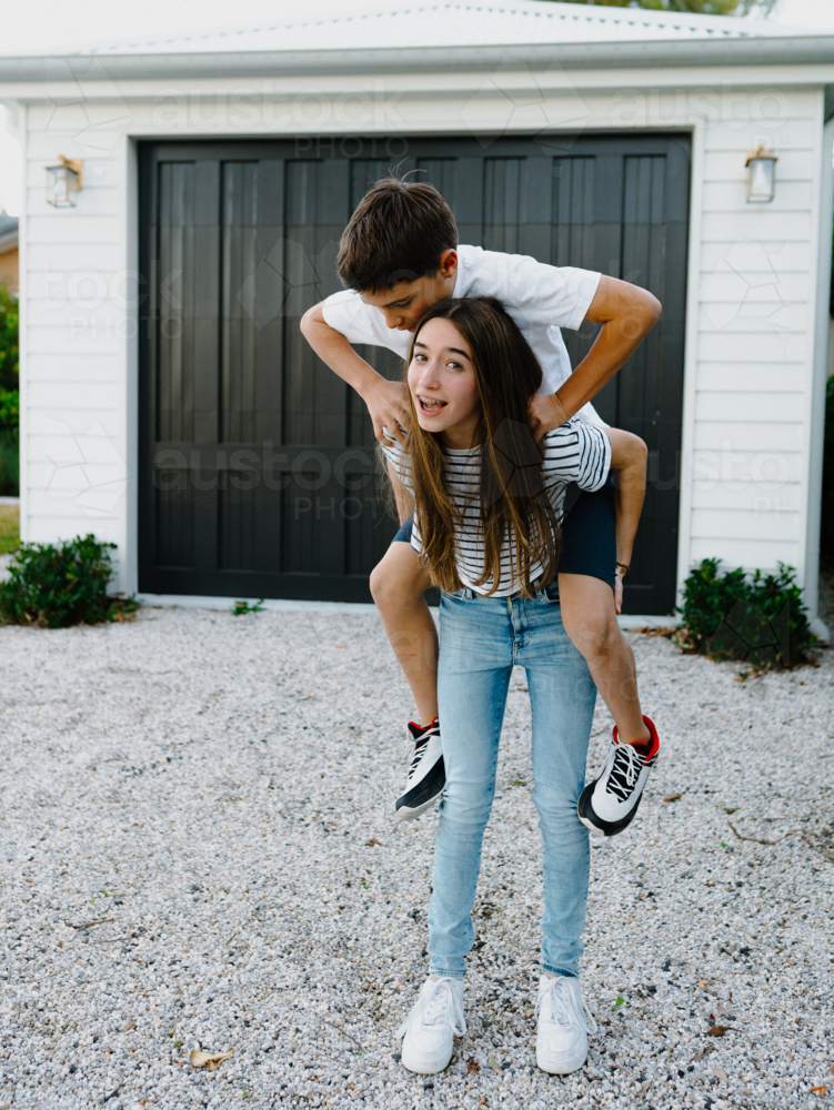 Teenage girl giving her brother a piggyback around their yard at home - Australian Stock Image