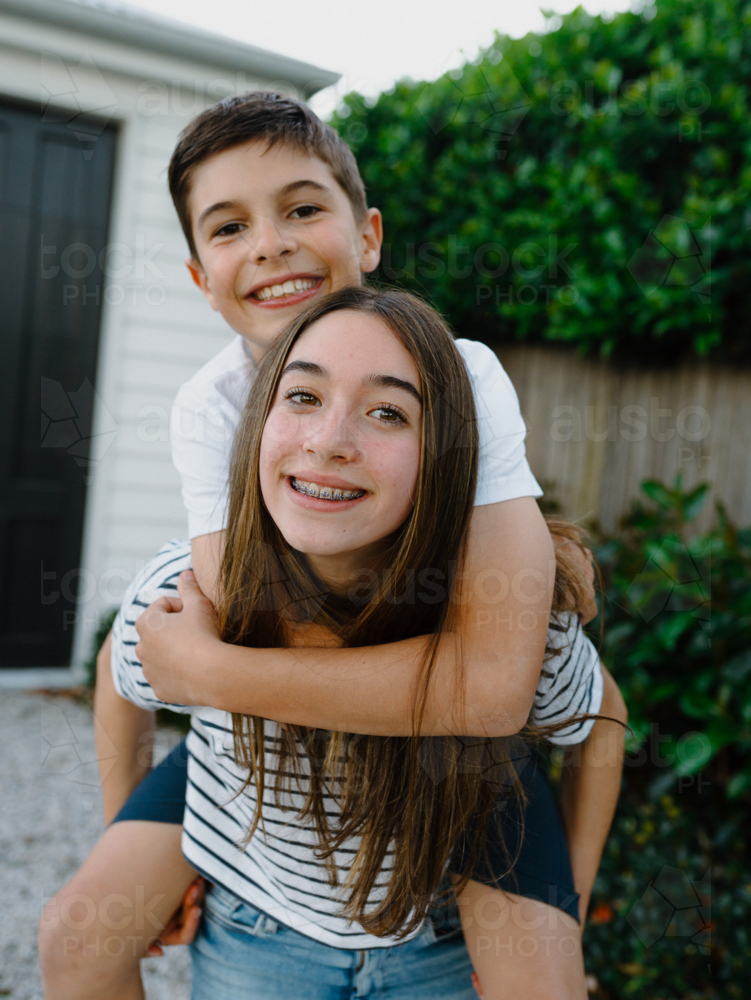 Teenage girl giving her brother a piggyback around their yard at home - Australian Stock Image