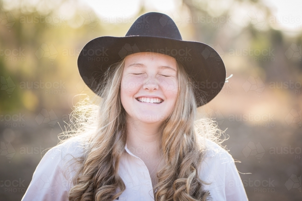 Teenage girl laughing with eyes closed wearing akubra hat on farm in drought - Australian Stock Image