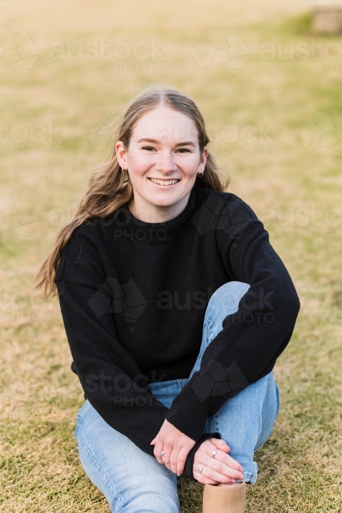 Teenage girl sitting on grass smiling with arm leaning on leg - Australian Stock Image