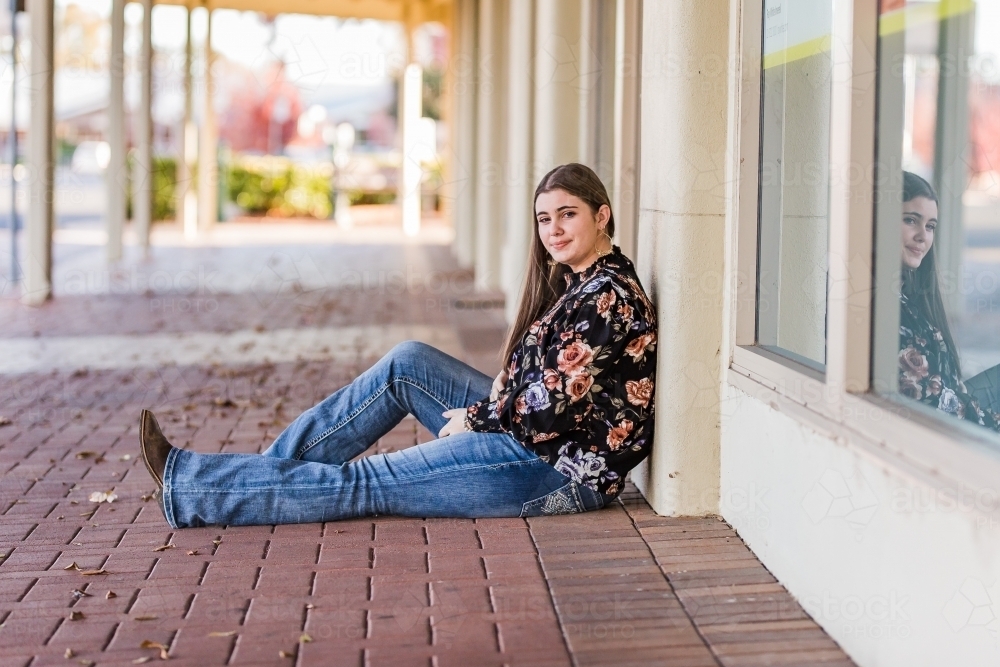 Teenager sitting on main street leaning against wall smiling - Australian Stock Image