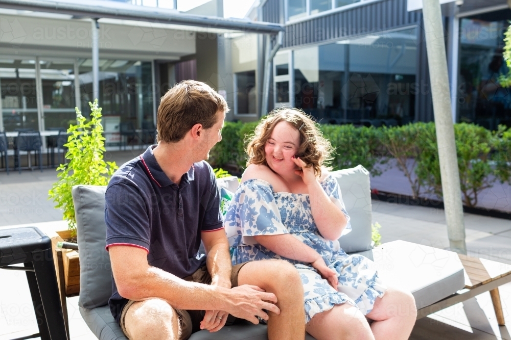 Teenager talking with young adult disability worker outside at café sitting on lounge together - Australian Stock Image