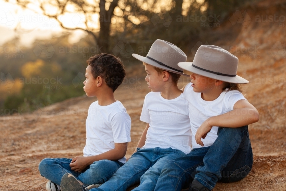 Three aboriginal kids sitting together in rural Australia looking out over landscape at sunset - Australian Stock Image