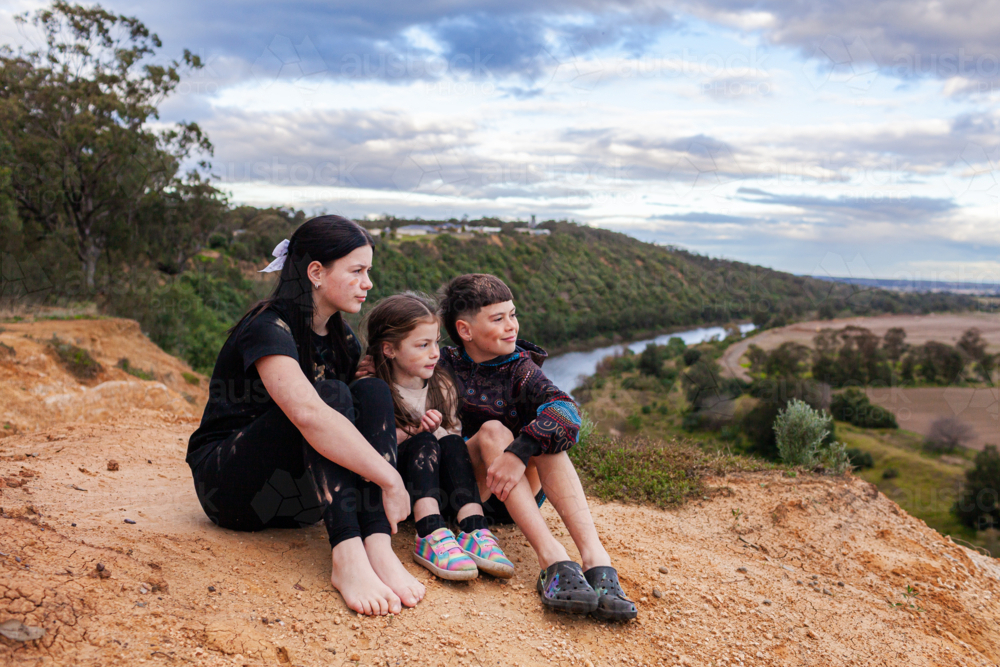 Three aboriginal siblings sisters and brother sitting together on country looking out over landscape - Australian Stock Image