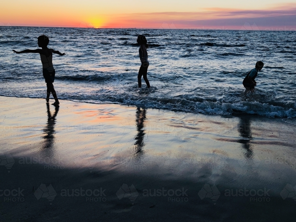 Three children playing on oceans edge with sun setting in background - Australian Stock Image