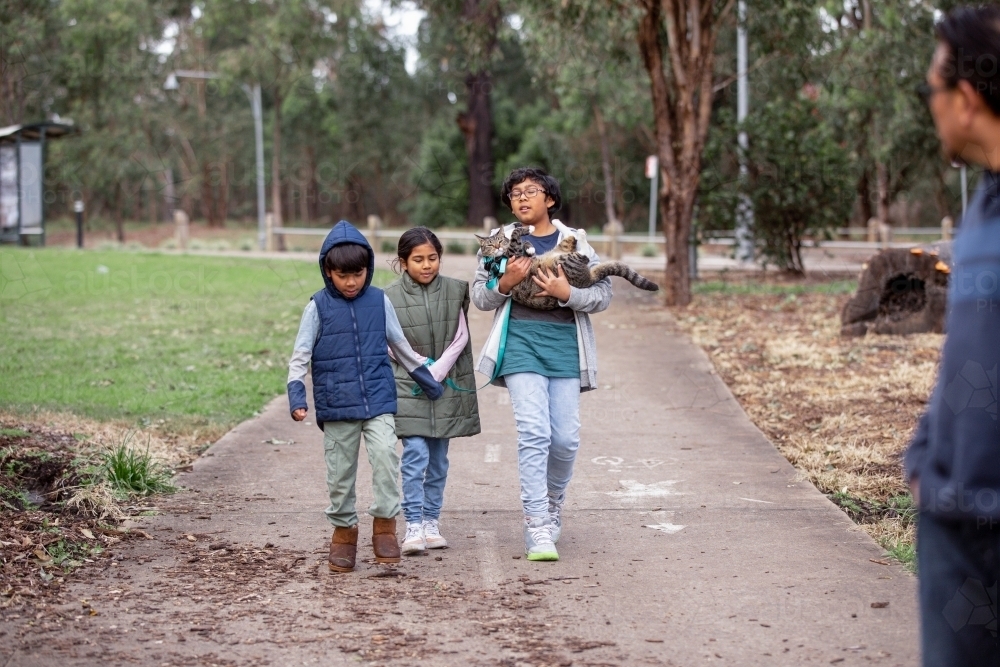 Three children with one boy carrying a cat on a footpath at park - Australian Stock Image