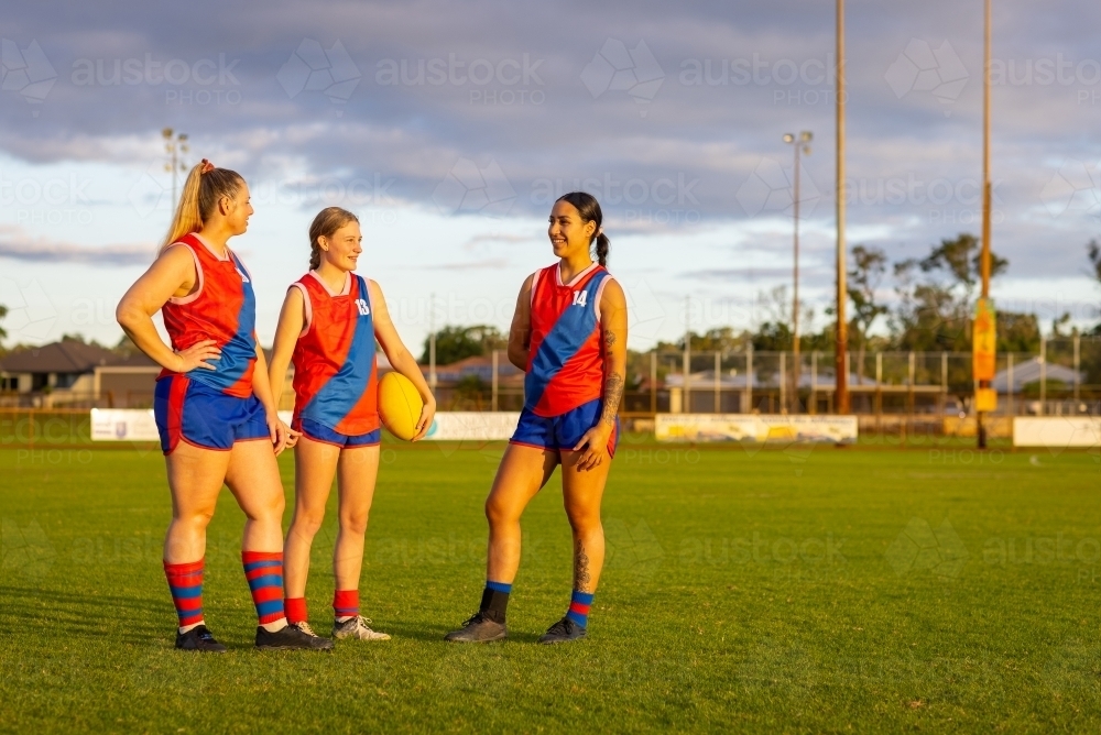 three female footballers standing on footy oval with football in late afternoon light - Australian Stock Image