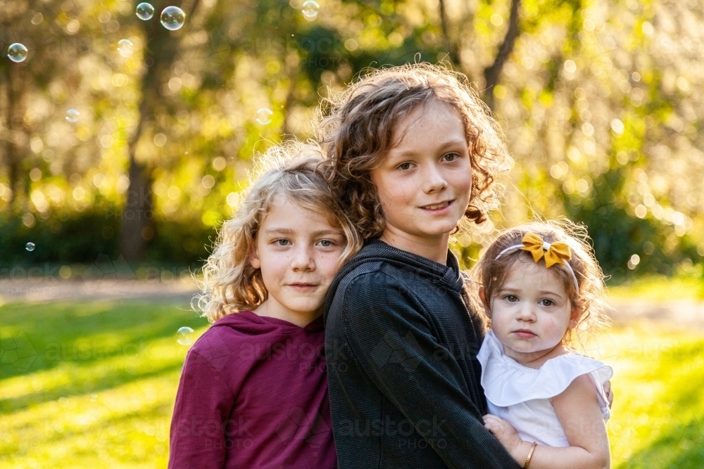 Three happy siblings with curly hair smiling at camera in park - Australian Stock Image
