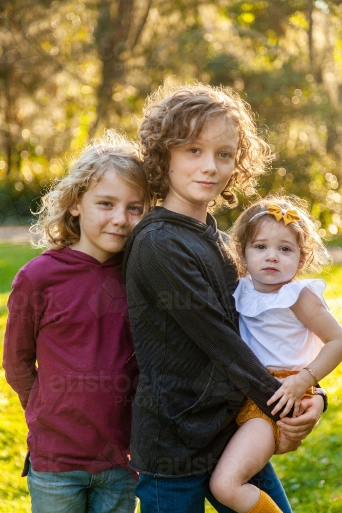 Three happy siblings with curly hair smiling at camera in park - Australian Stock Image