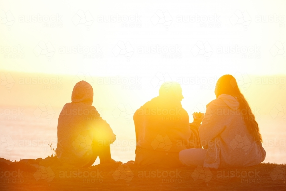 Three people sitting by the beach at sunset. - Australian Stock Image