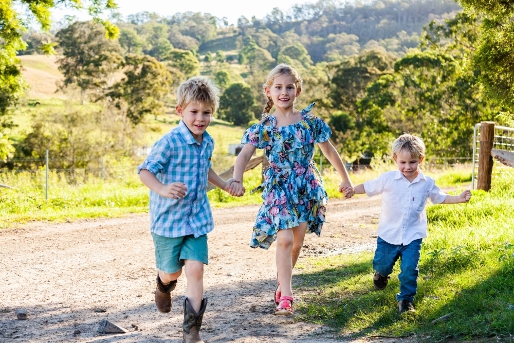 Three siblings running down the farm driveway, playing a game together - Australian Stock Image