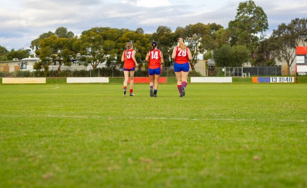 three young female footballers jogging across football oval - Australian Stock Image