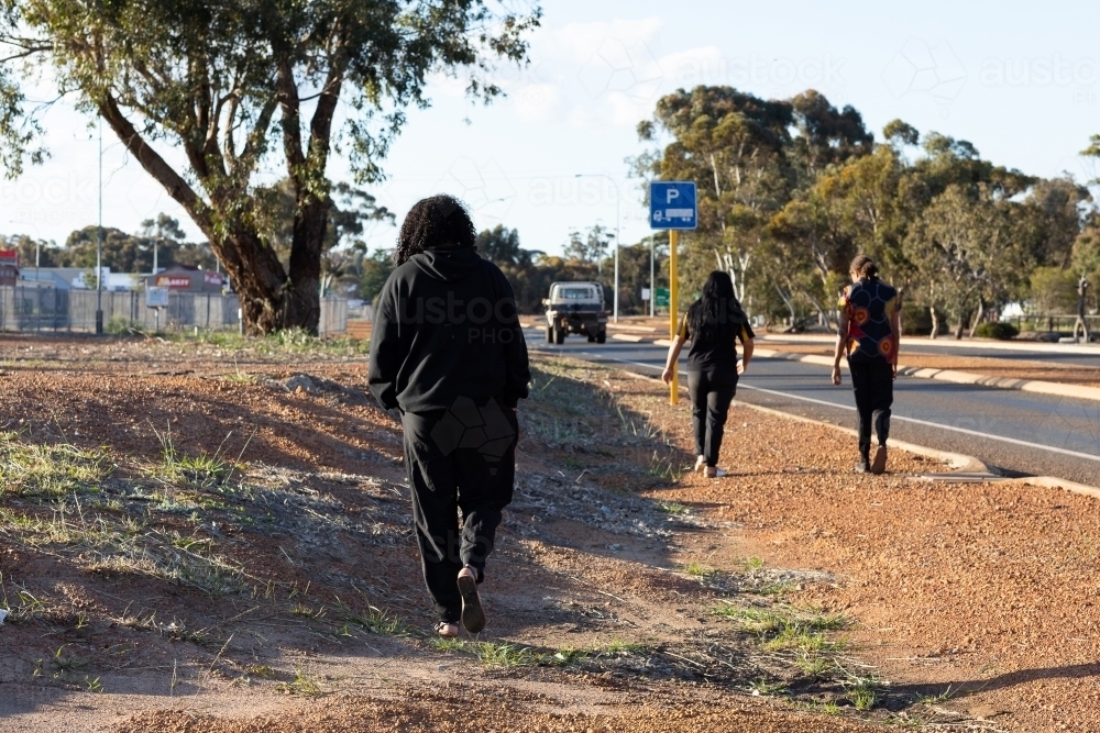 three young people dressed in black walking along side of highway in regional town - Australian Stock Image