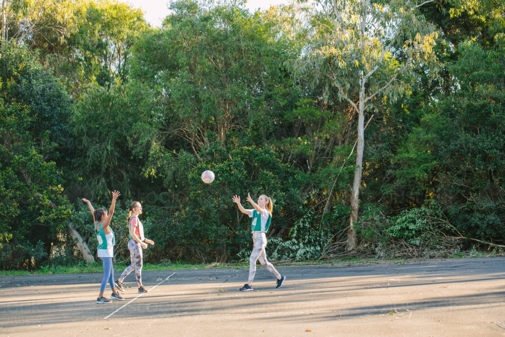 three young women with one throwing a net ball in mid air on a sunny day - Australian Stock Image