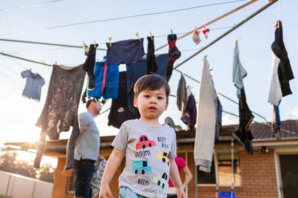 Toddler boy in backyard playing while parents get in washing from hills hoist line - Australian Stock Image