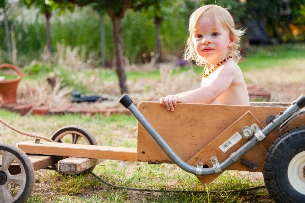 Toddler boy sitting in billy cart in backyard smiling - Australian Stock Image