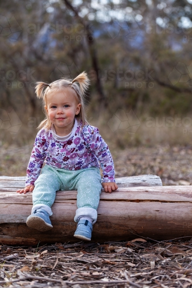 Toddler child sitting on log in bushland camping area - Australian Stock Image