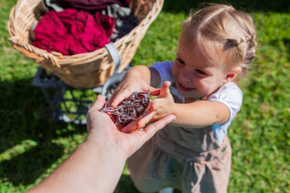 Toddler girl handing pegs to mum to help with hanging out washing in backyard - Australian Stock Image