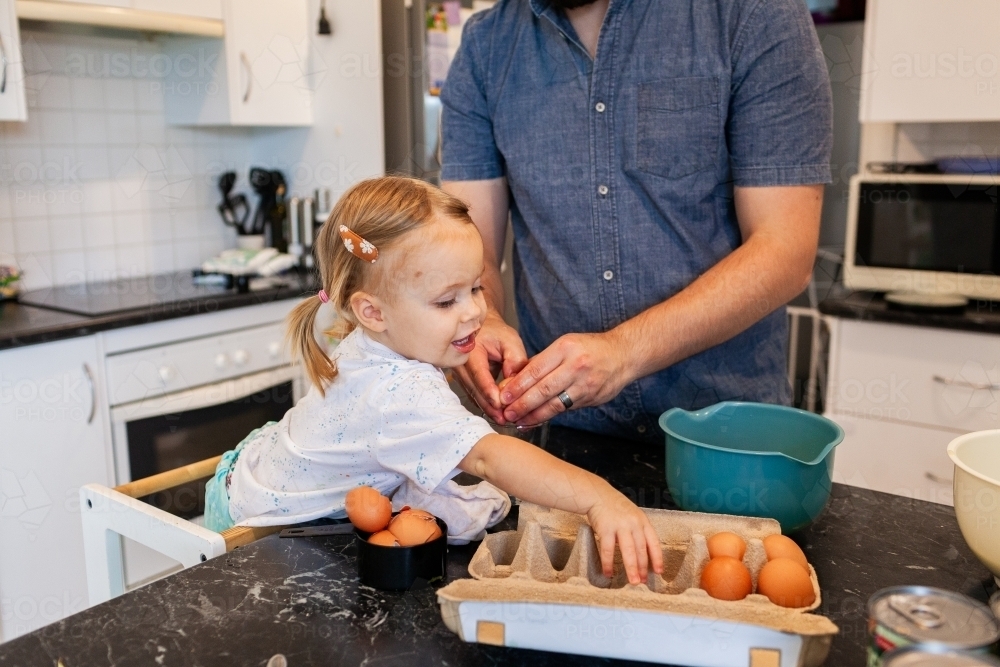 Toddler girl helping crack eggs for baking a cake with her dad in kitchen - Australian Stock Image