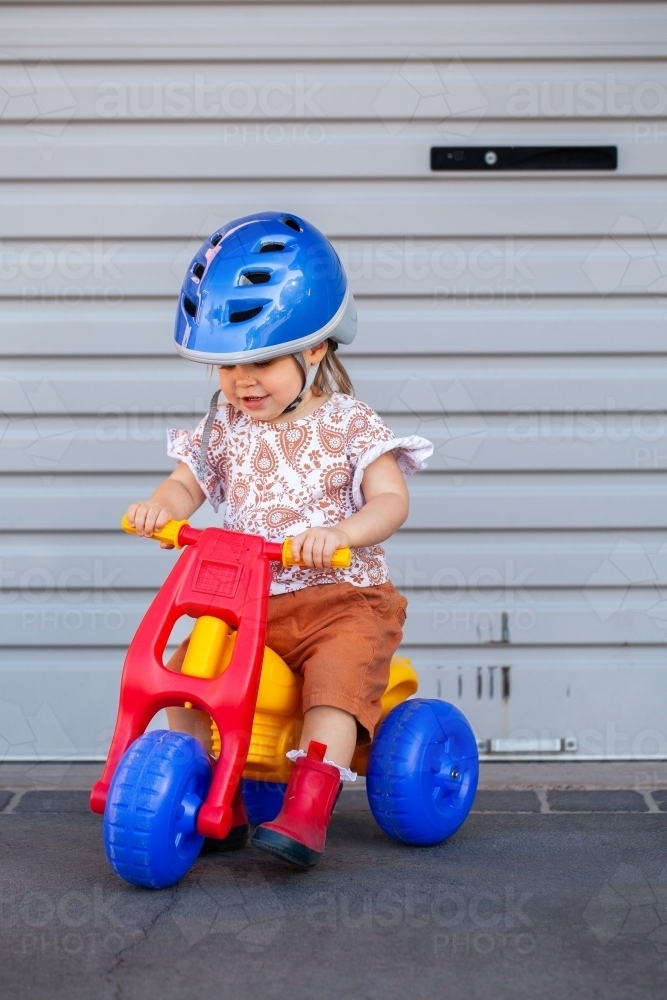 Toddler girl riding colourful tricycle outside garage - Australian Stock Image