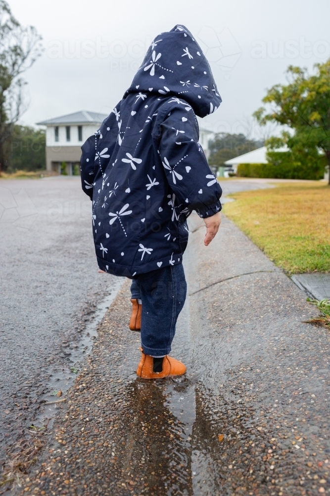 Toddler girl wearing raincoat splashing in gutter at roadside - Australian Stock Image