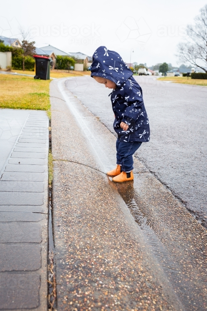 Toddler girl wearing raincoat splashing in gutter at roadside - Australian Stock Image