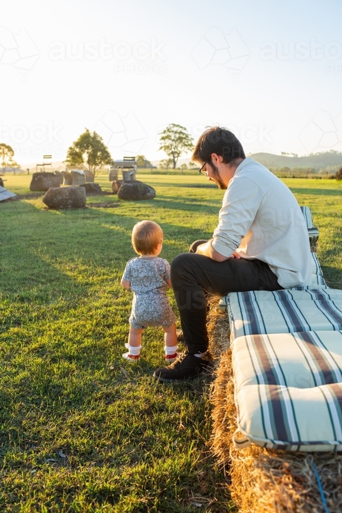 Toddler with dad outside at campsite bonfire night sitting on hay bale seat - Australian Stock Image