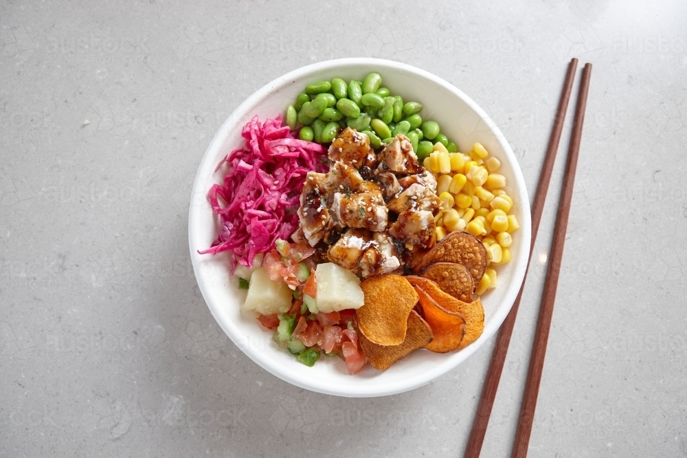 top shot of a bowl of chicken vegetable salad with wooden chopsticks on top of a dirty white surface - Australian Stock Image